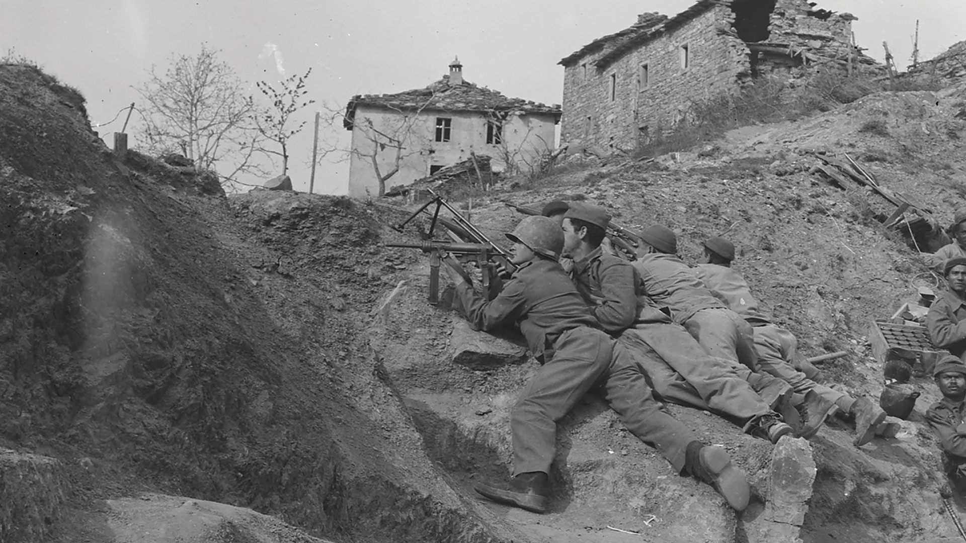 Black and white photo of Brazilian soldiers in a trench aiming a machine gun near a damaged building during World War II.