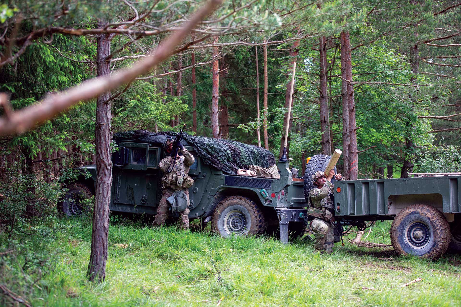 Soldiers in woodland beside a camouflaged Humvee and trailer, one soldier aiming a rifle upward