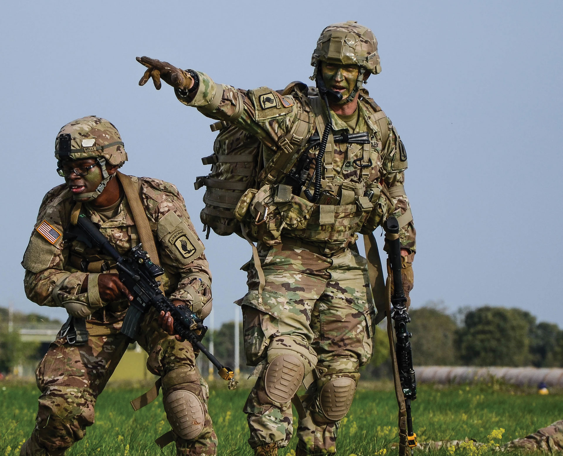 Two Soldiers holding rifles stand in a field with face paint and wearing camo gear.
