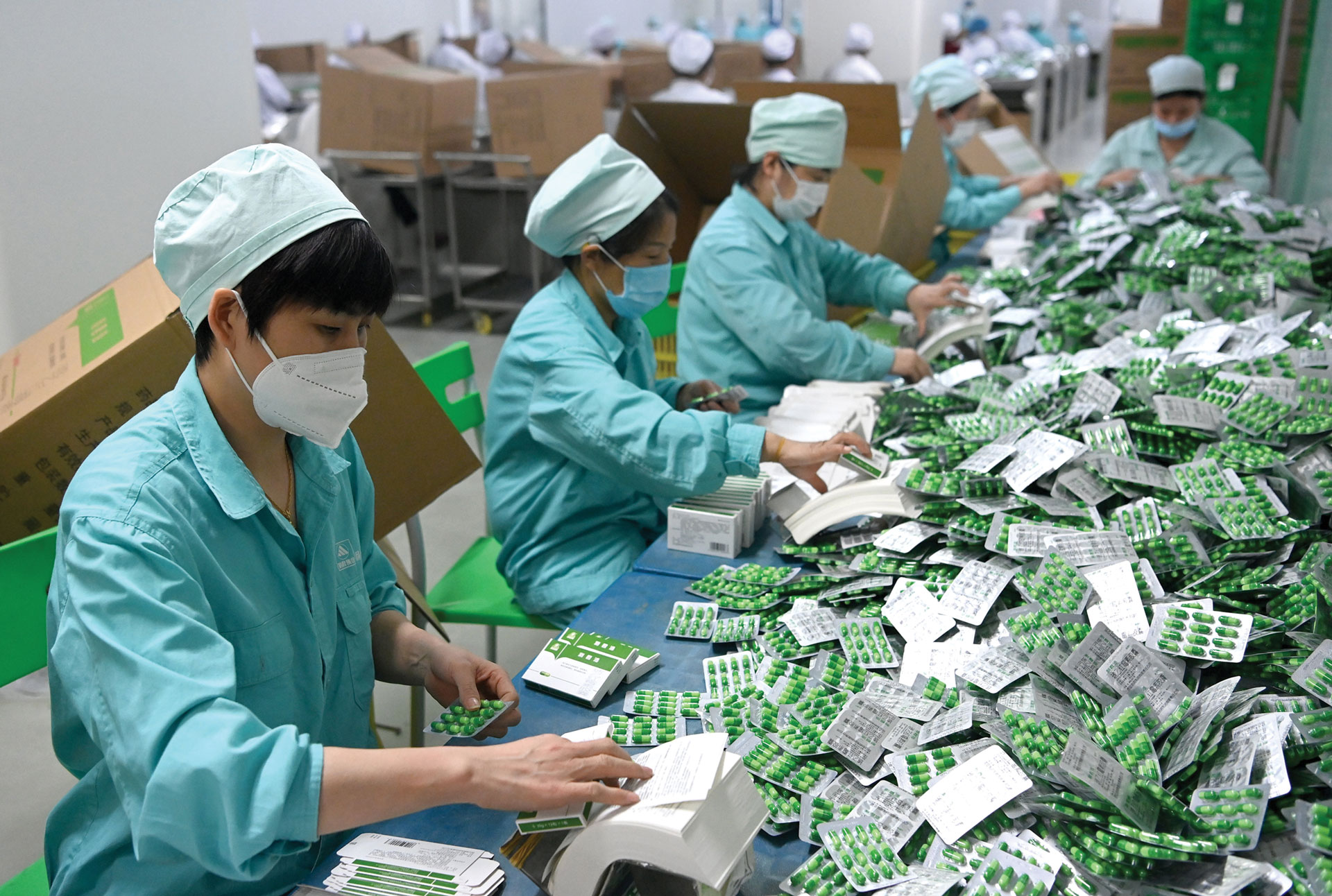 Workers wearing masks and teal uniforms package green and white blister-pack pills at a pharmaceutical manufacturing facility.
