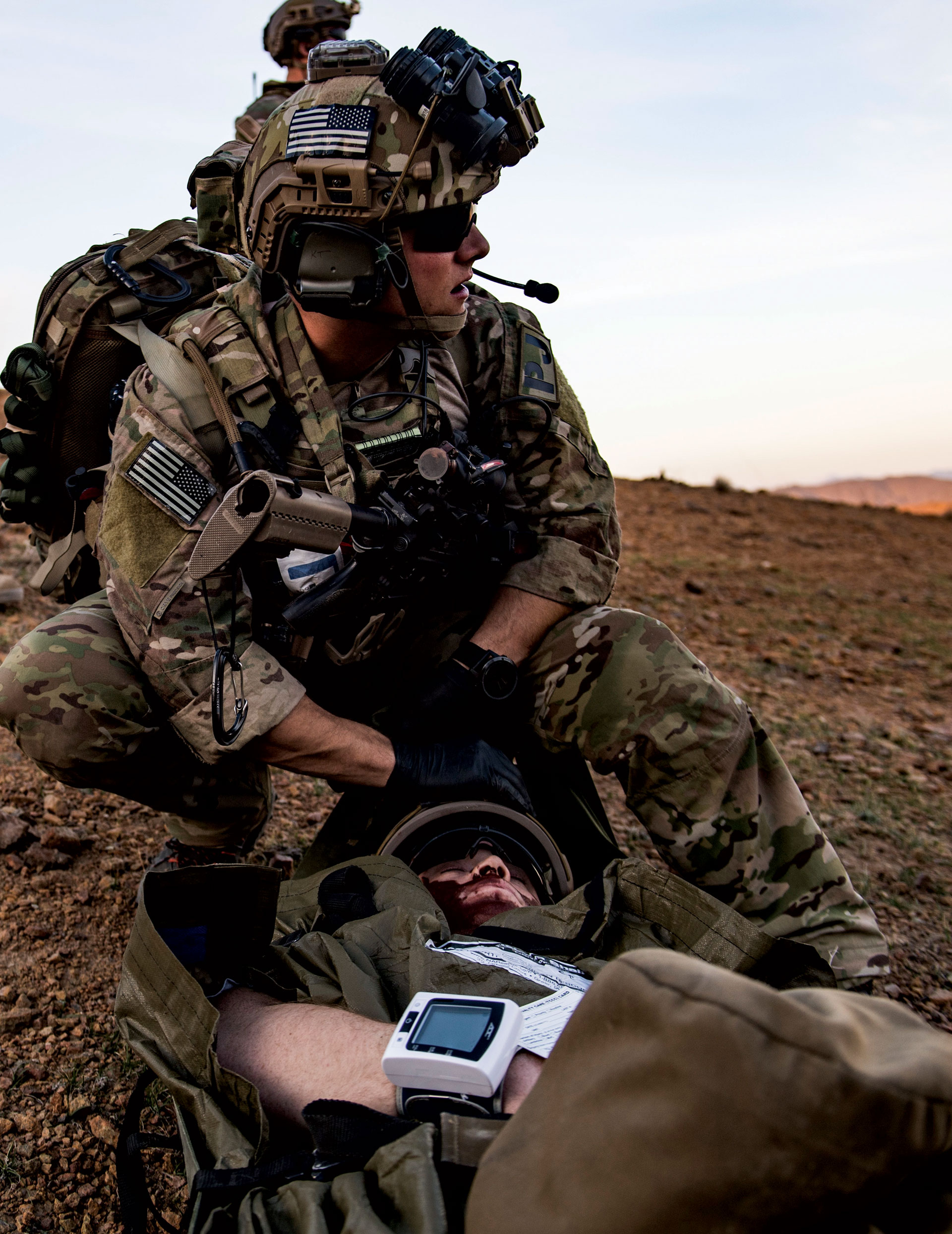 Military personnel providing aid to a simulated wounded soldier in a rocky outdoor environment.
