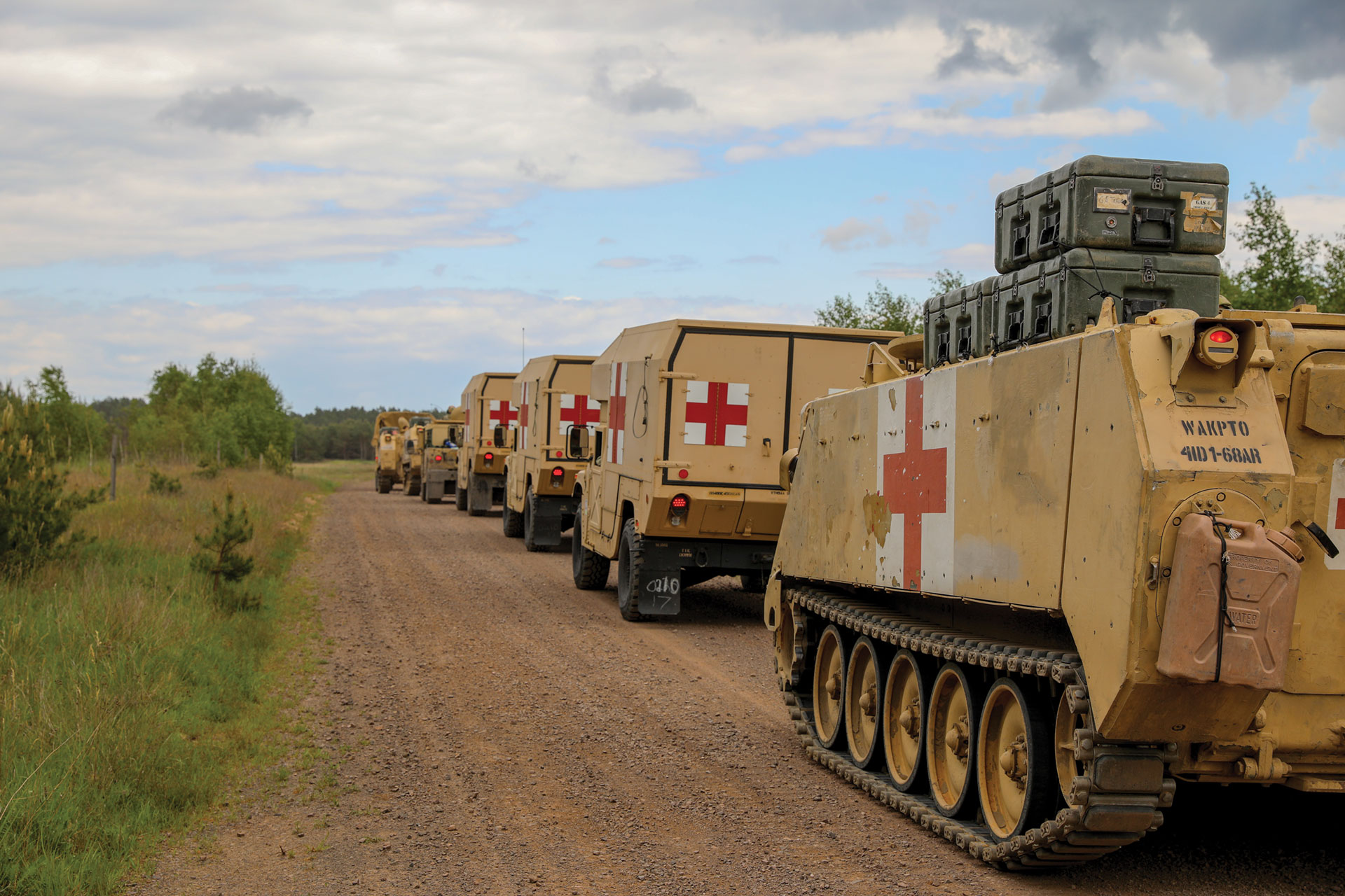 Line of tan-colored military ambulances with red cross symbols traveling on a dirt road in a wooded area under a partly cloudy sky.