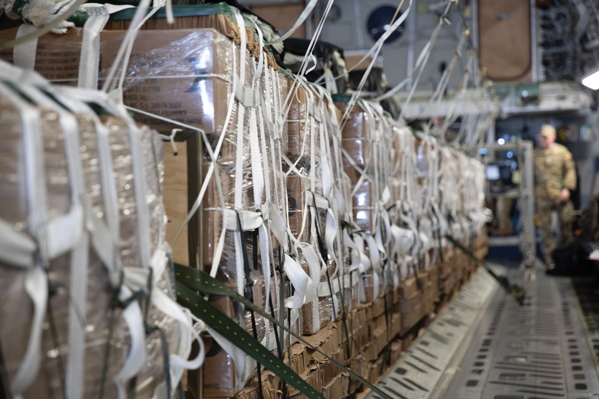 Rows of tightly strapped cargo pallets inside a military transport aircraft, with a soldier visible in the background.