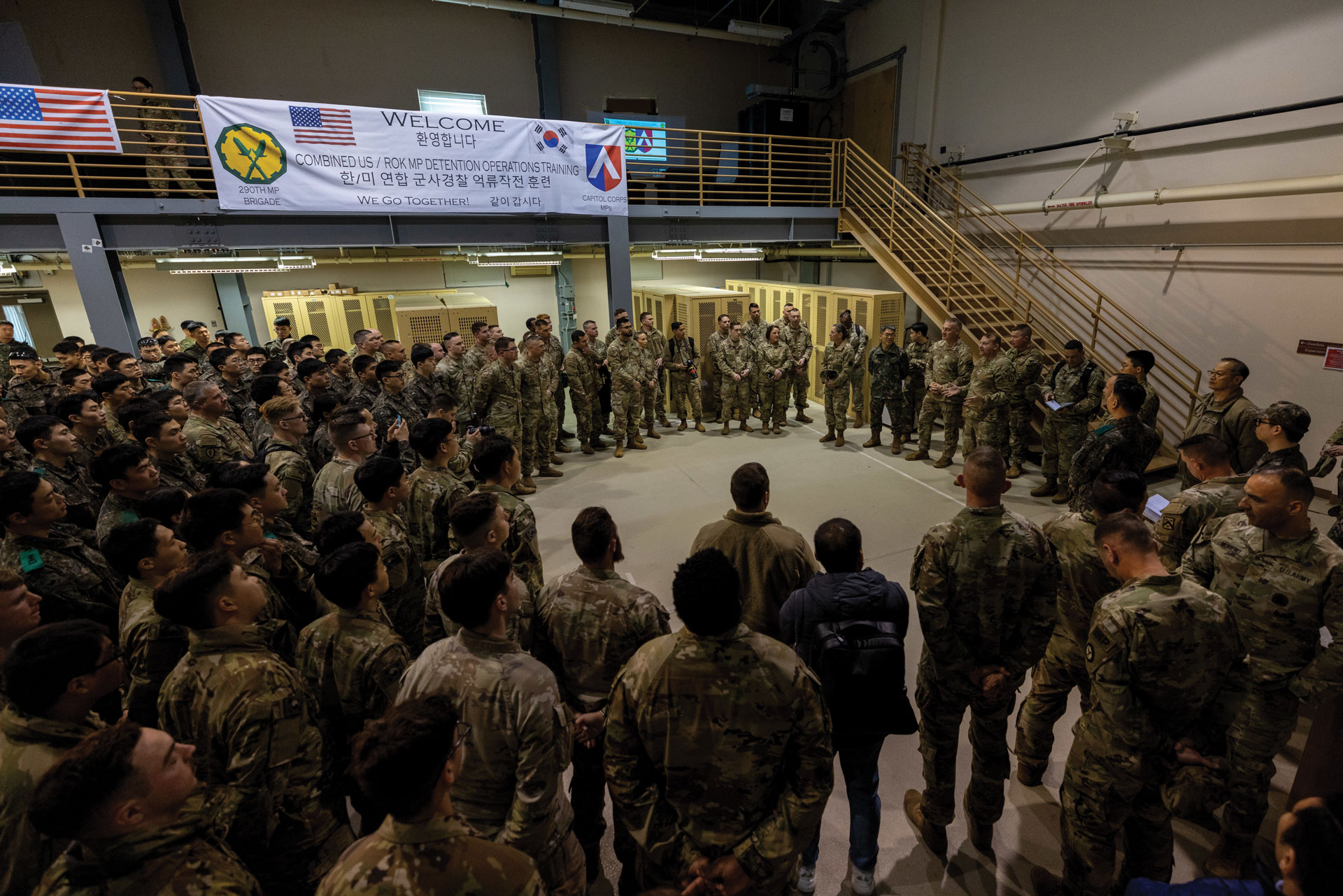 Lt. Gen. Jody J. Daniels speaks to a A large group of military personnel gathered in a semicircle for a briefing or meeting indoors, with a banner overhead welcoming them to a joint detention operations training.