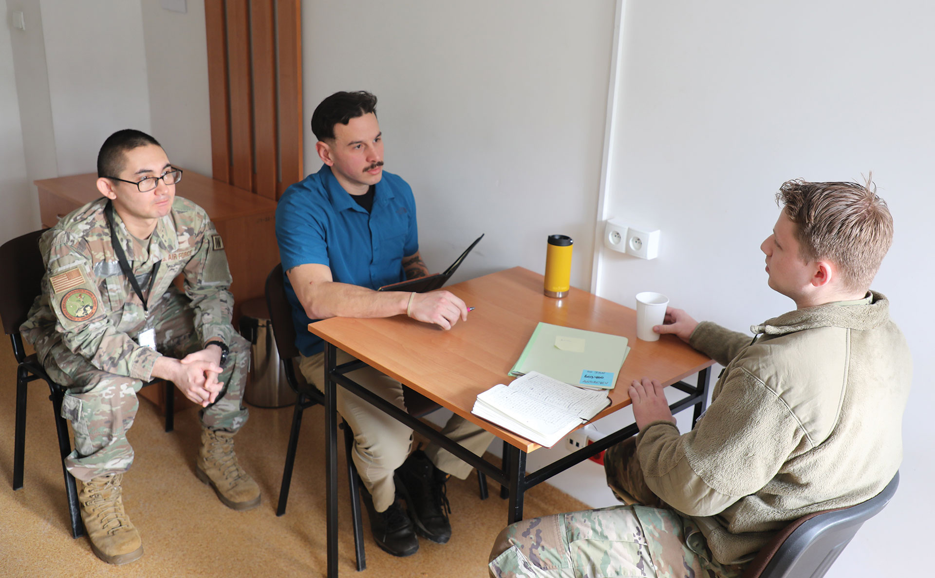 Three men seated at a small table indoors, conducting an interview or debriefing session with another soldier. Documents and a coffee cup are on the table.