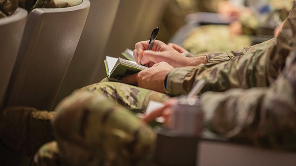 Detalhe da mão de um militar enquanto escreve em um caderno verde sentado em um auditório com outros militares.