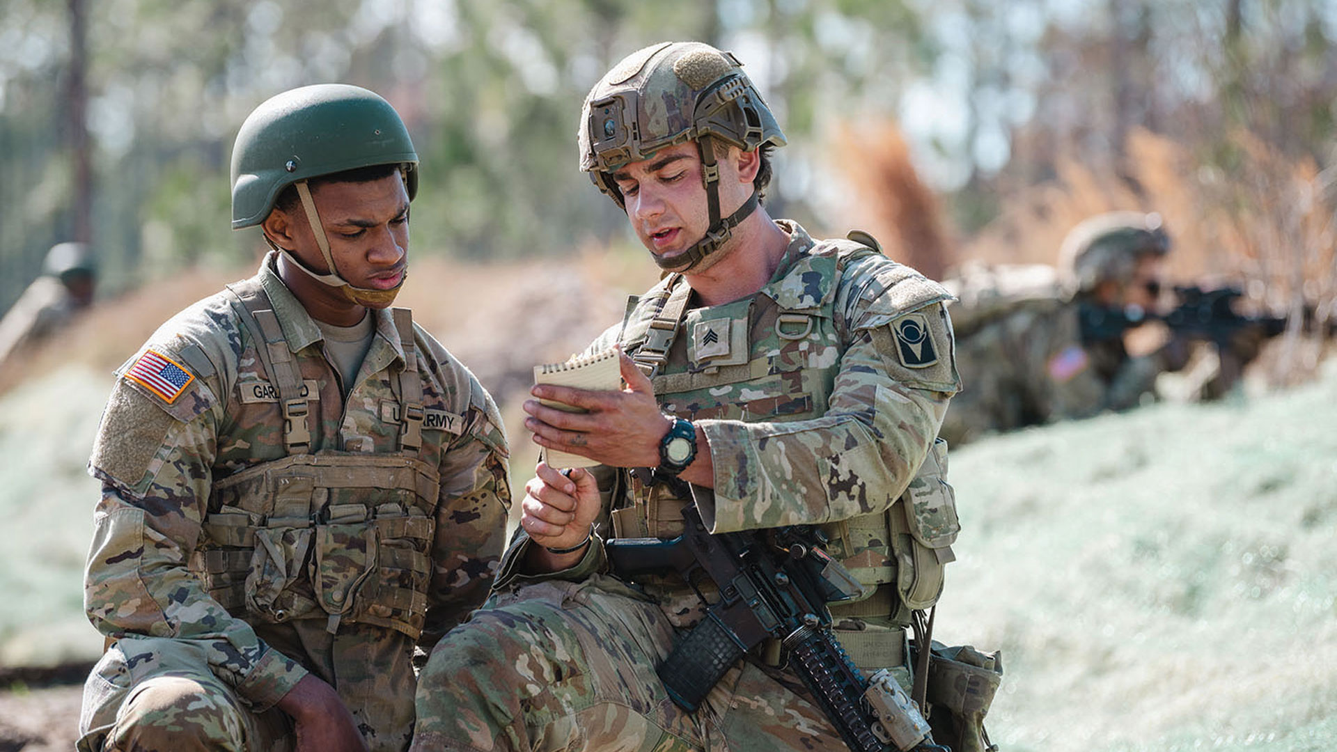 Dois soldados em camuflagem revisando anotações em um caderno durante exercício ao ar livre