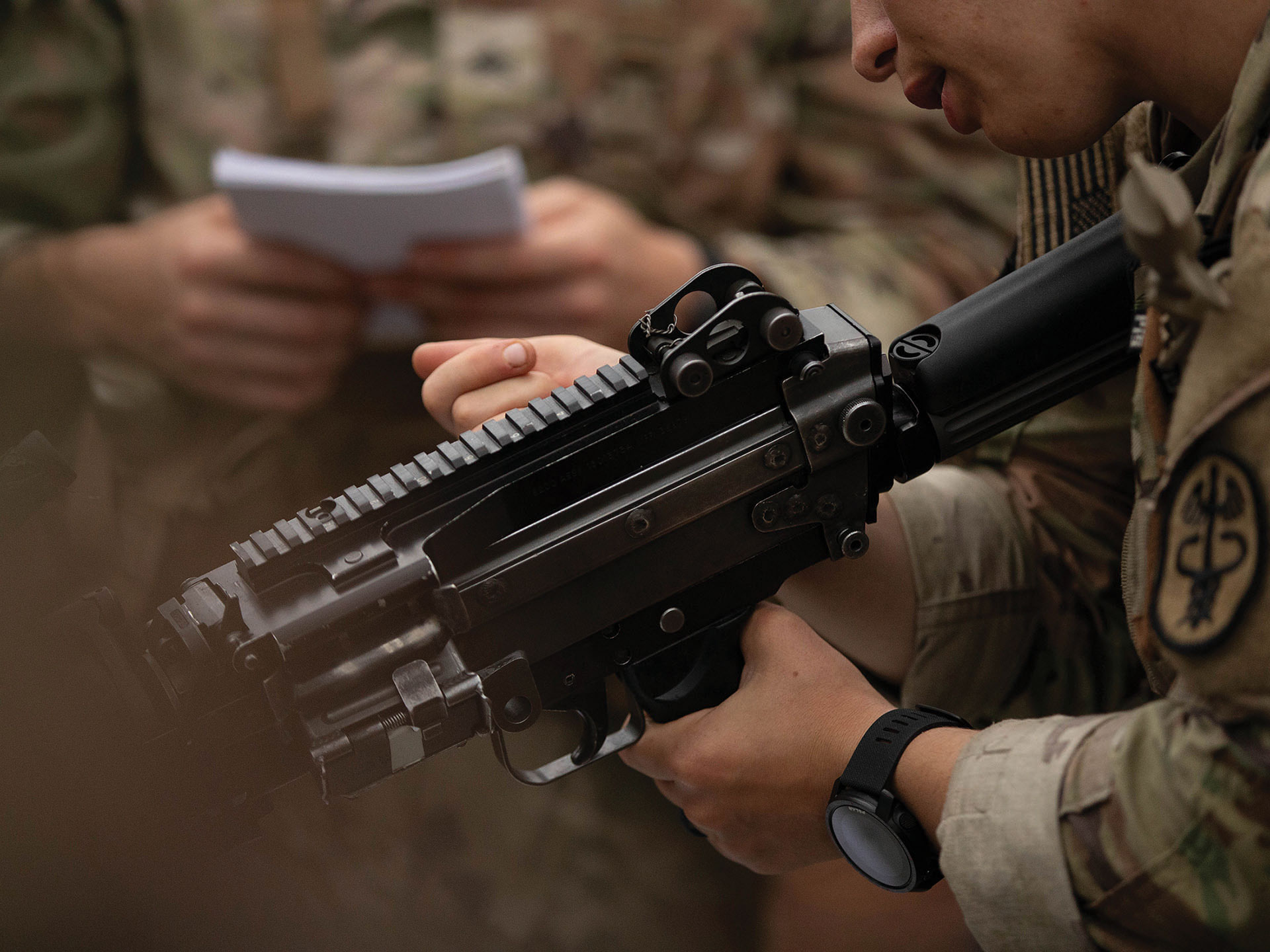 Soldado manuseando metralhadora enquanto outro soldado lê anotações em caderno ao fundo
