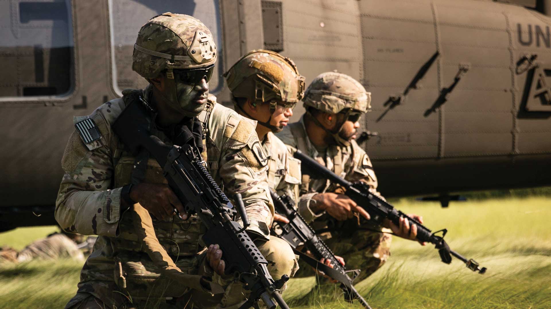 Tres soldados completamente equipados y portando sus armas están en cuclillas en un terreno cubierto de pasto.