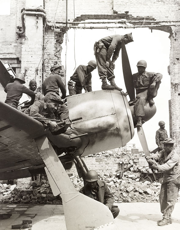 Soldiers of the 4046th Quartermaster Truck Company inspect and remove pieces from a grounded fighter plane found within a bombed-out plant