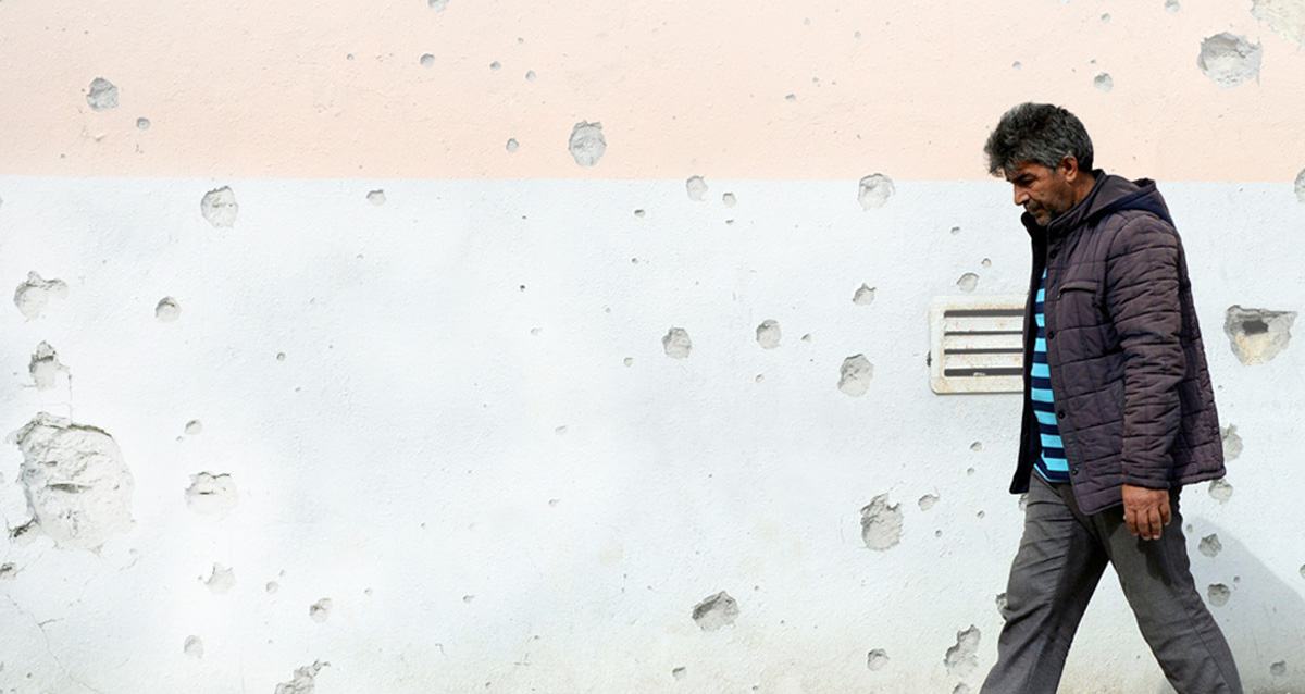 Photo of a man walking next to wall with bullet holes