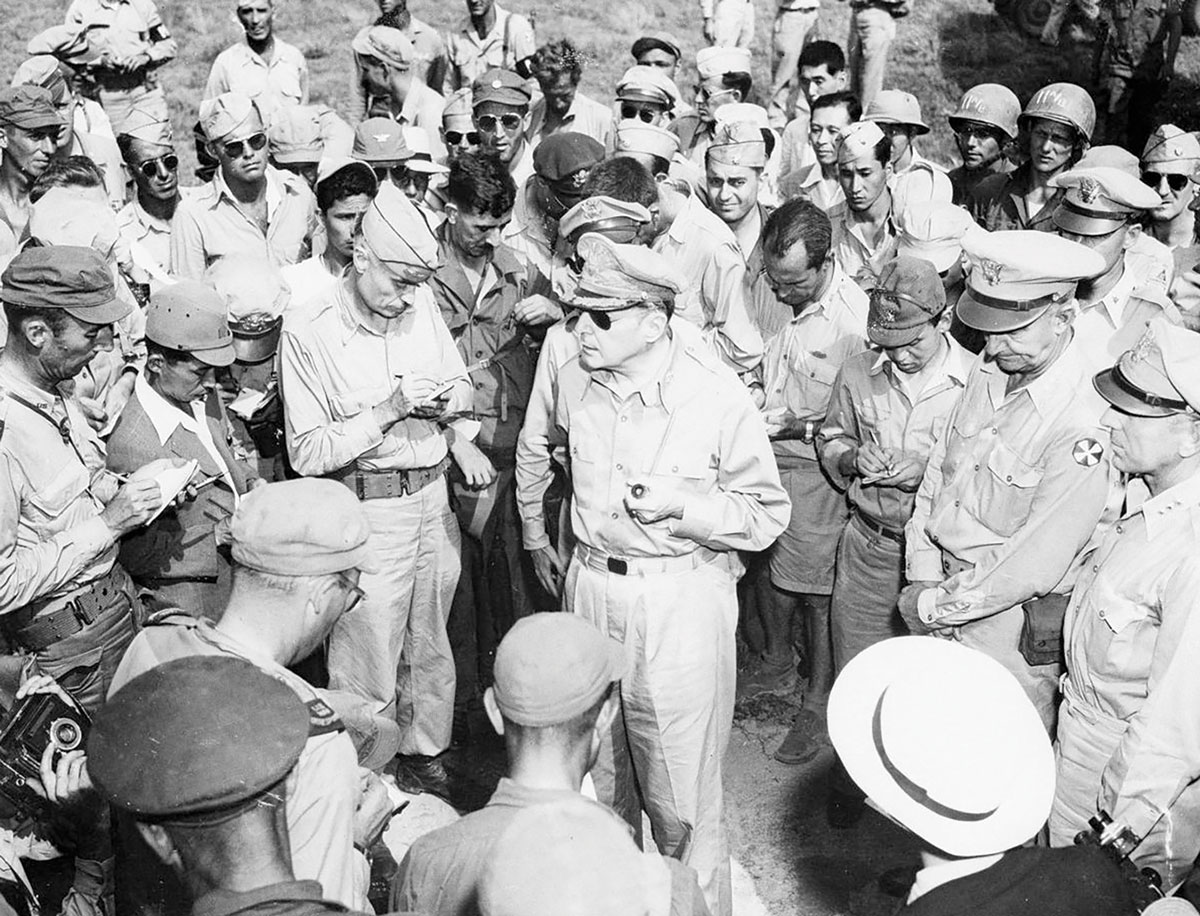 O General Douglas MacArthur (centro), acompanhado do Gen Div Robert L. Eichelberger (à direita, com as mãos entrelaçadas na frente do corpo), responde a perguntas de repórteres, logo após sua chegada ao Campo de Pouso de Atsugi, no Japão, 30 Ago 1945. (Foto do Arquivo Nacional dos EUA)