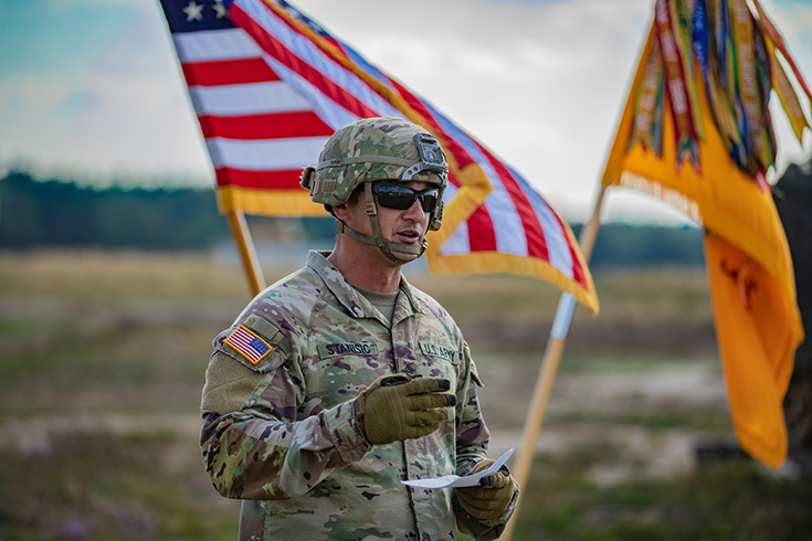 Outside, a Soldier in helmet, sunglasses, and gloves holds a sheet of paper before him as he speaks. An American flag and a yellow flag wave in the wind behind him.