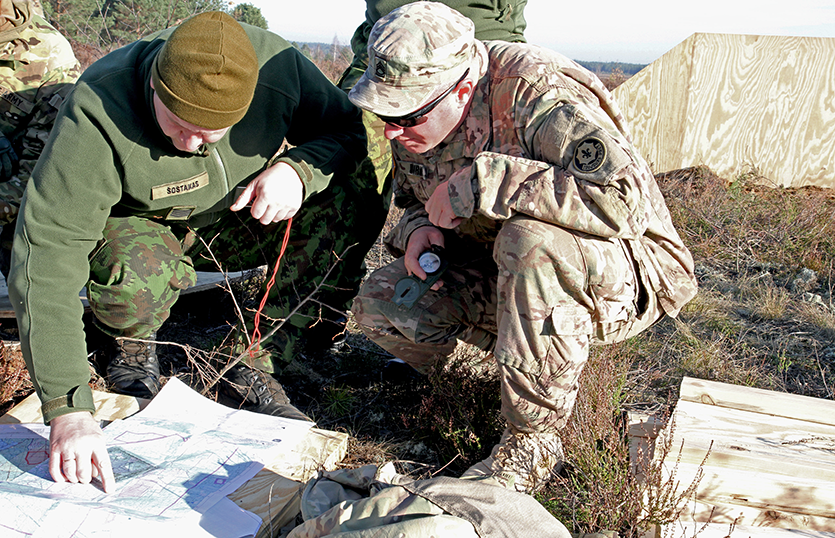 A pair of Soldiers kneel to study a map unfolded on the ground. One wears a green top, brown cap, and brown-and-green camo pants. The other wears a standard uniform of the U.S. Army.