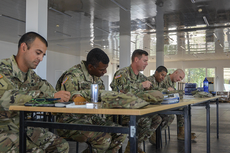Five bareheaded Soldiers sit at a long table in a room with windows looking out upon green trees. Each stares down and writes with pen on material on the tabletop before them.
