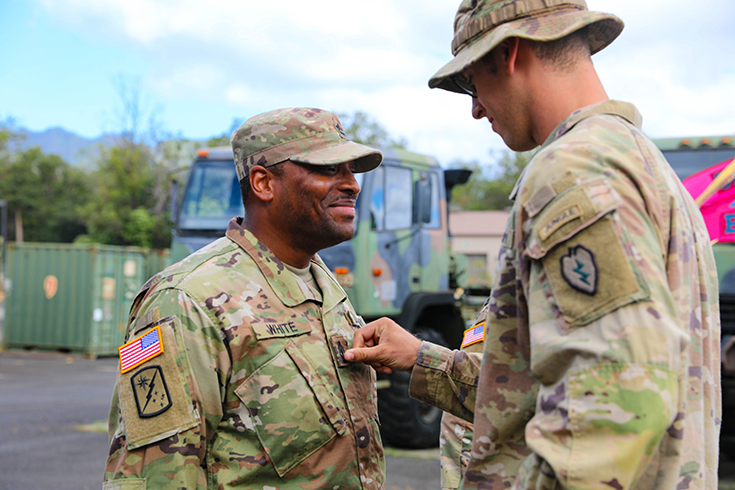 Two Soldiers stand outside on a bright day, one pinning a badge to the chest of the other Soldier before him.