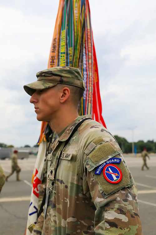 A vertical photograph shows a Soldier standing outside in camouflaged uniform and cap. A patch on his upper arm reads “Honor Guard.” At his side are what appear to be multicolored flag streamers. The gaze of the Soldier leads the viewer’s eye toward the left edge of the photograph. This image is the second in a series of five accompanying the article.