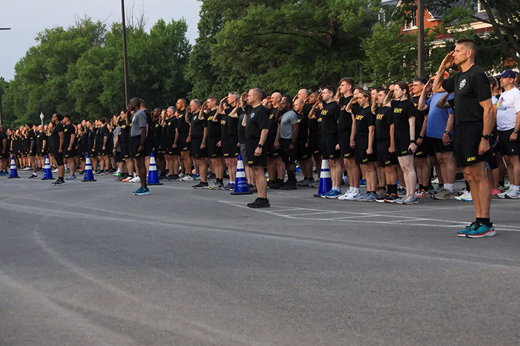 A horizontal photograph shows a crowd of men and women in black shirts and shorts, “Army” emblazoned in yellow on their chests, standing at salute while lining the side of a road behind a row of blue traffic cones. This image is the third in a series of five accompanying the article.
