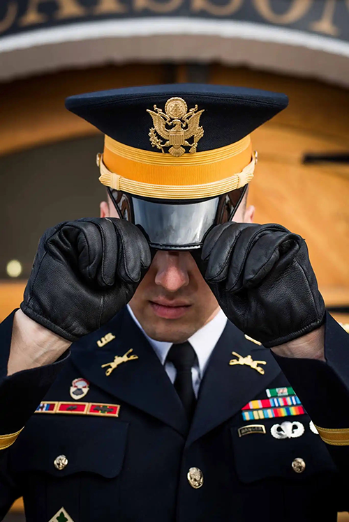 A vertical photograph shows a Soldier in a navy-blue dress uniform and black gloves, pulling on the brim of a cap with a black visor. This image is the fifth in a series of five accompanying the article.