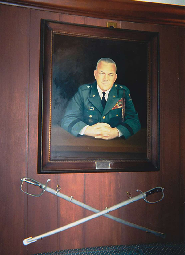 A vertical photograph shows a painted portrait of a uniformed Soldier with closely shaved white hair, sitting at a desk. The frame of this portrait hangs on a wood-paneled wall, above a pair of crossed sabers mounted below it. Keen observers will note a resemblance between the painted image in this color photograph and the black-and-white photograph inset in the article’s lead image. This image is the twelfth in a series of 12 accompanying the article.