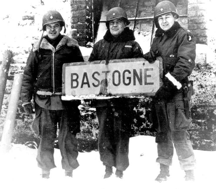 A horizontal photograph shows two uniformed Soldiers in helmets holding a battered sign reading “Bastogne” before them. The image is black and white. Snow dusts the bottom of the sign, and the men wear gloves and coats with thick collars. A stone wall rises behind them, and a third man is partially visible at the edge of the photograph’s frame. This image is the fourth in a series of 12 accompanying the article.