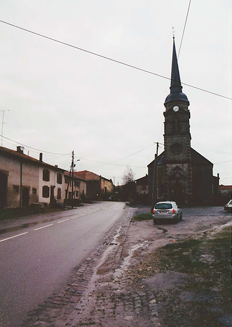 A vertical photograph shows a street lined by low, two-story buildings on one side and a tall, spired building with a clock on its face on the other. A sports utility vehicle is parked before the latter, its back lights bright red, as if a driver within is pressing on the brakes or has activated the vehicle’s headlights in anticipation of nightfall. This image is the sixth in a series of 12 accompanying the article.
