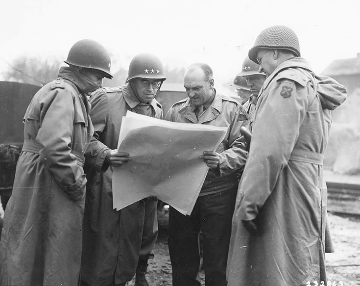 A horizontal photograph shows five Soldiers huddled together outside, looking at long, folio-sized sheets of paper held by two of the men. The image is black and white. All but one of the men wear helmets. The helmets whose fronts are visible to the camera’s lens bear three stars above the brim. This image is the seventh in a series of 12 accompanying the article.