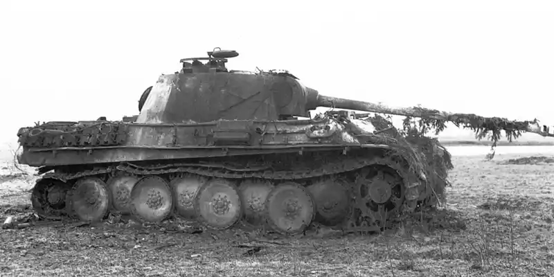 A horizontal photograph shows a dilapidated tank, with vegetation growing on its gun and between its worn track and battered wheels. The image is black and white. This image is the eighth in a series of 12 accompanying the article.