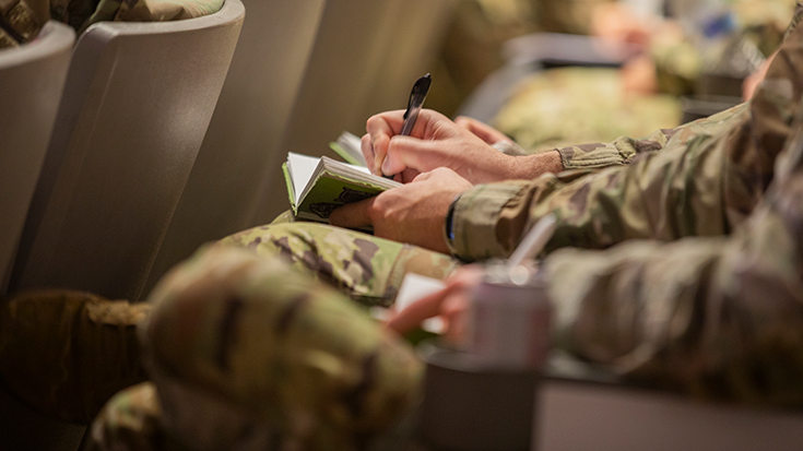In a photograph, one sees the hands of a Soldier using a pen to write in a notebook. One can also make out the backs of a row of seats before the writer, suggesting the setting of the shot is a classroom auditorium or a theater where a presentation is taking place.