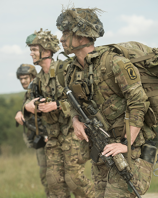 A vertical photograph shows a trio of helmeted Soldiers standing with rifles held before them and packs on their backs. Beyond them, a clear sky meets the top of a grassy hill. The Soldiers face forward, looking before them, their gaze leading the viewer’s eye toward the left edge of the photograph.  This image is the second in a series of three accompanying the article.