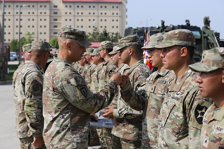 A horizontal photograph shows a line of Soldiers standing shoulder-to-shoulder outside, military vehicles behind them, and a beige, multistory building rising into a blue sky in the background, beyond green trees and lampposts. Standing before the line of Soldiers are three other Soldiers, one of whom exchanges a fist bump with one of the men standing before him. This image is the third in a series of three accompanying the article.