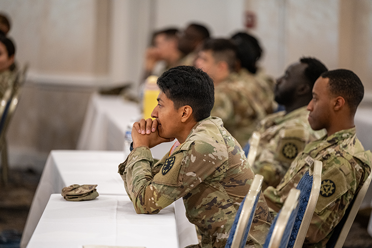 This lead image for the article is a photograph of a Soldier at a table topped with white linen. She leans forward as she listens intently, elbows on the surface before her, and her chin resting upon her thumbs as she stares forward contemplatively. Beyond her, other Soldiers sit beside her at other tables extending to a wall in the background, in what appears to be a banquet-style meeting room. This image is the first in a series of three accompanying the article.