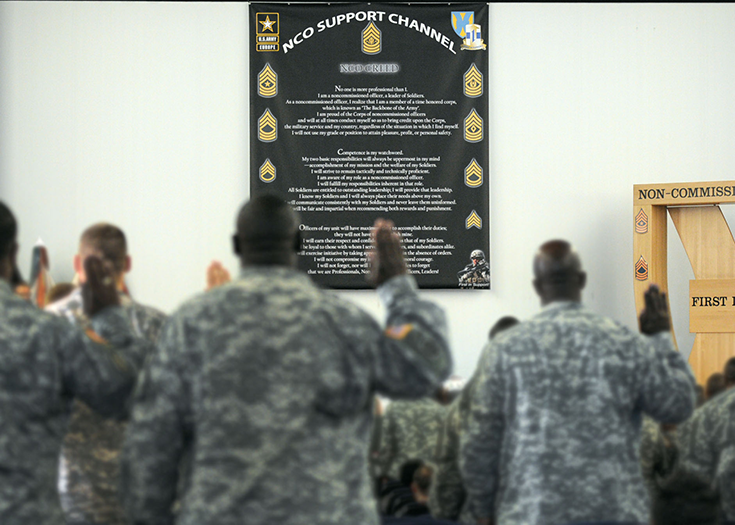 Soldiers standing side by side in a room with white walls raise their right hands, as if to take an oath. On the wall before them hangs a poster filled with text below the legend “NCO Support Channel.”