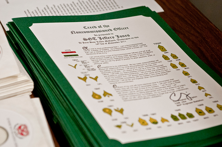 A stack of green diploma-like documents lies atop a desktop. While most of the text on the topmost paper is too small to make out in the photograph, the first lines of it — in a larger font — read, “Creed of the Noncommissioned Officer Is presented to …”