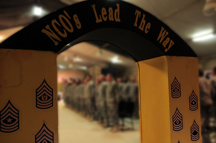 A horizontal photo shows a yellow arch in what looks like an auditorium. In the background, one distinguishes the blurred figures of Soldiers. They stand, as if participating in a ceremony. Atop the arch, writing spells out “NCO’s Lead the Way.”