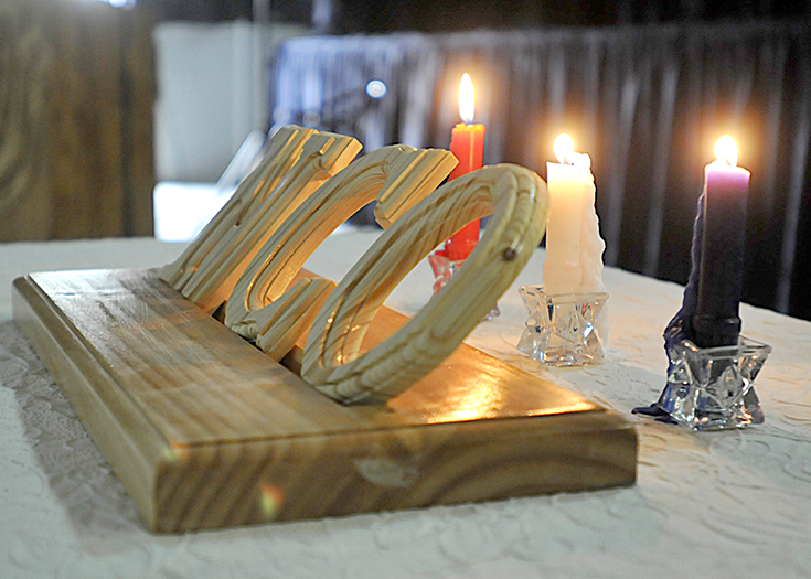 A photograph shows a wooden plaque with the letters “NCO” atop a wooden base. It rests on a tabletop before three lit candles.