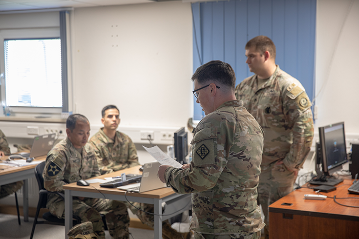 A horizontal photograph shows a bespectacled Soldier standing in a classroom-like setting, where other Soldiers sit at tables with laptops before them. The standing Soldier looks down at printed sheets of paper held in hand while addressing the room, and another Soldier stands near him, looking at the seated Soldiers before them. This image is the second in a series of six accompanying the article.