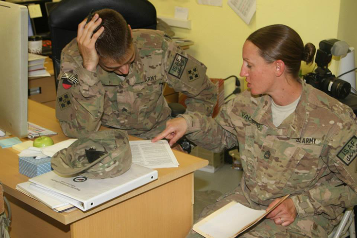 A horizontal photograph shows a pair of Soldiers sitting at a desktop bearing a computer, a green apple, a box of tissues, yellow and blue sticky note pads, and a stack of paperwork. One Soldier points to the bottom of a printed form, while the other leans over it with a pen in hand. This image is the fourth in a series of six accompanying the article.