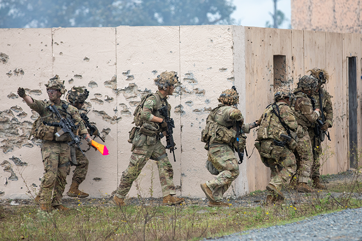 A horizontal photograph shows a line of helmeted Soldiers, rifle in hand, rounding the corner of a low building. One Soldier holds a bright yellow-and-magenta cloth in one hand while gesturing with the other, suggesting the men may be participating in a training exercise. This image is the fifth in a series of six accompanying the article.