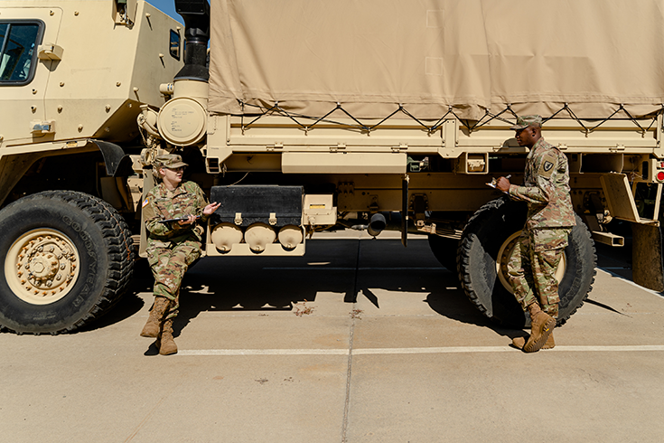 A horizontal photograph shows a capped Soldier sitting on the side of a military vehicle, her boots planted on the sand-colored pavement before her. She gestures with one hand and holds a clipboard in the other, as she speaks to a Soldier standing some 10 feet away from her. This second Soldier also wears a cap, and he puts a pen to the clipboard he holds before him. One of the vehicle’s tires rises behind him to the height of his abdomen, and behind his head rises the bottom of the sand-colored canvas covering the vehicle’s cargo, which rises beyond the top of the photograph’s frame. This image is the sixth in a series of six accompanying the article.