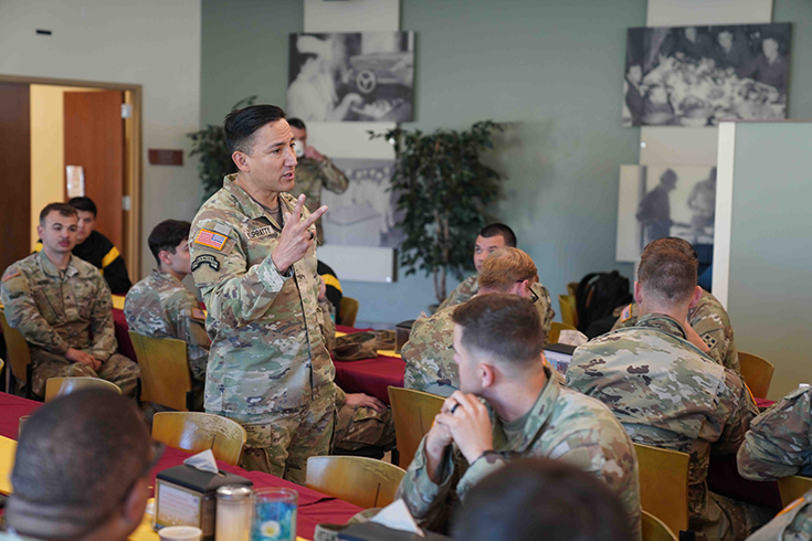 This lead image for the article is a photograph of a Soldier holding up two fingers as he speaks, standing between two long dining tables where other Soldiers sit and listen attentively. In the background is a green wall bearing black-and-white images of Soldiers, which meets a white wall with a door that opens onto a hallway with yellow walls. This image is the first in a series of six accompanying the article.