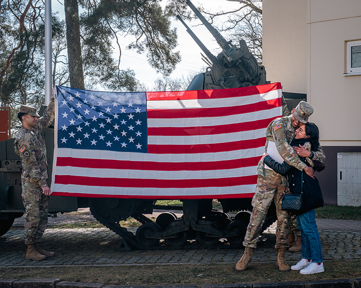A Soldier hugs a woman in jeans and sneakers outside of a beige building. Directly behind them, two other Soldiers hold up an American flag roughly the length and height of a subcompact car. It obscures from the camera’s lens the treaded artillery vehicle directly behind it and the piney trees that reach into the bright sky beyond.