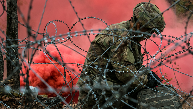 Through a snarl of barbed wire, a camera captures the image of a helmeted Soldier sitting on a leaf-strewn ground. He turns his gaze down toward his lap as if in defeat. In the background, salmon-colored smoke spews and rises, obscuring the greenery beyond its thick plumes.