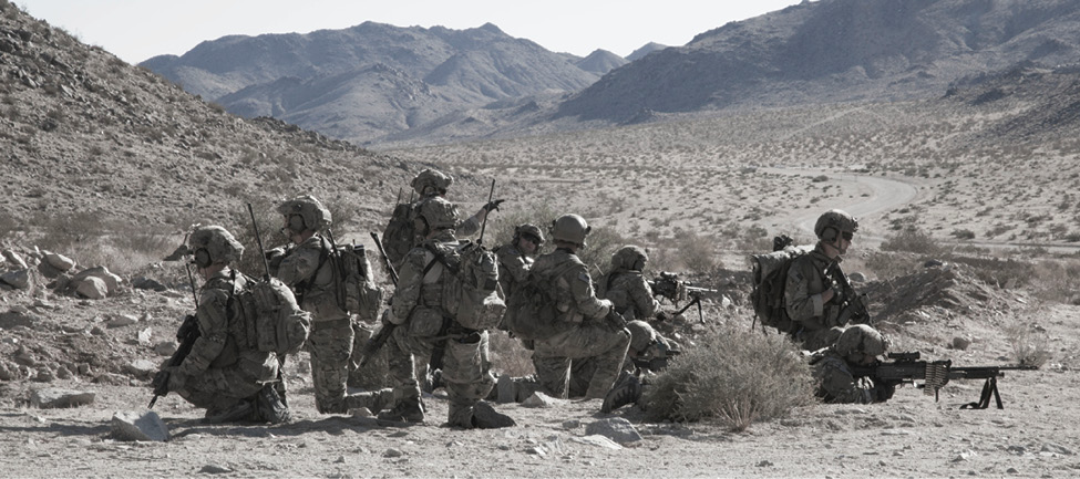 A horizontal photograph shows some 10 Soldiers crouching or lying on a rocky plain, some with rifles and scopes before them. The sky above is a bright white, and the direct light turns the men and landscape around them into shades of gray and black. Only distant mountains in the background add color to the image, rising blue at the far horizon.