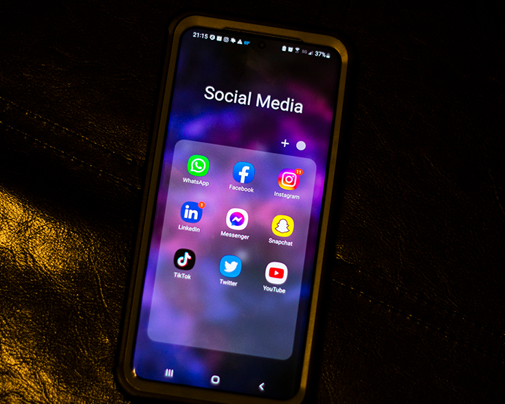 A close-up photograph of a smartphone shows a screen labeled “Social Media,” containing nine icons for apps including Facebook, Instagram, LinkedIn, and more.