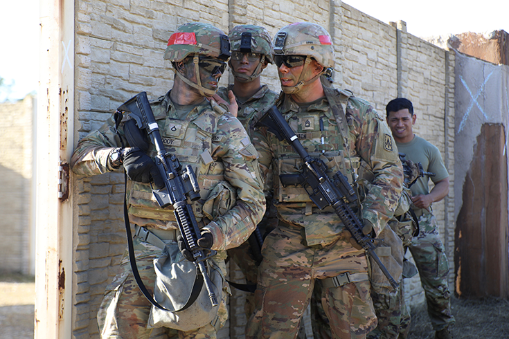 Three helmeted Soldiers in facepaint stand before a wall outdoors, rifles held before them, speaking together as if strategizing their next movements.