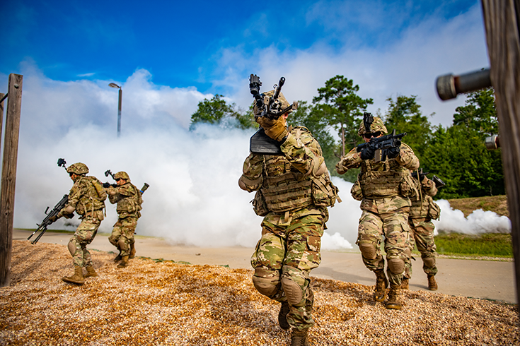 Five helmeted Soldiers wearing vests and kneepads advance with rifles drawn as plumes of white smoke billow near the road the squad crosses.