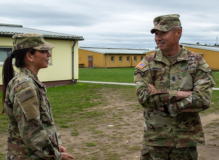 Two Soldiers in caps stand outside on an overcast day, speaking together while smiling outside a series of one-story buildings in the background.