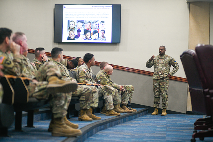 A Soldier stands near the front of what appears to be an auditorium, addressing a crowd of other Soldiers who listen attentively from the rows of seats before him.