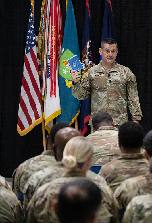 A Soldier standing before a row of flags and black curtains holds a blue book in hand. In the foreground of the photo, one sees the tops of other Soldiers’ heads, suggesting the man is addressing an auditorium from a stage. While it’s difficult to make out the man’s name on the chest of his uniform, many will recognize him as Michael Weimer, the sergeant major of the Army. 